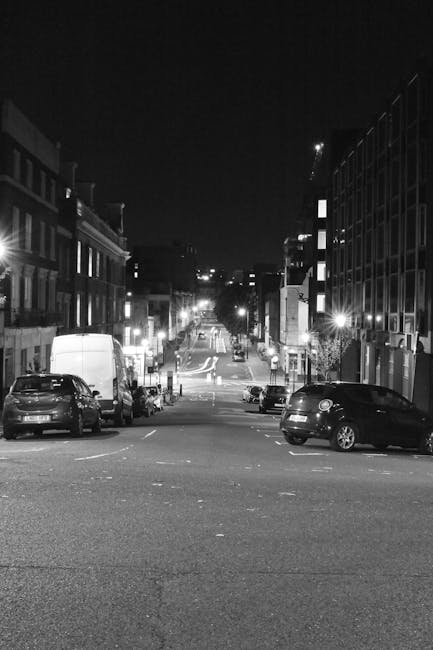 A black-and-white photograph of a narrow urban street at night, with parked cars lining both sides of the pavement. The street slopes slightly downward with some vehicles visible further along, and il